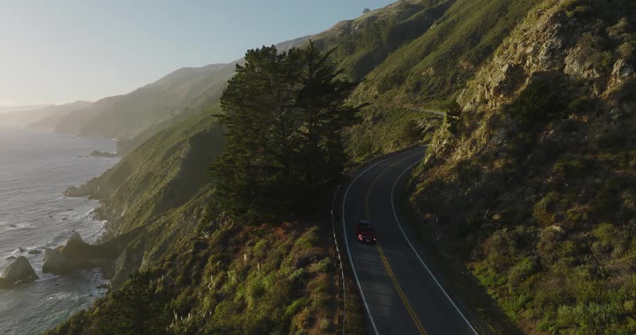 Red vehicle moves along highway hugging mountainside near foggy ocean