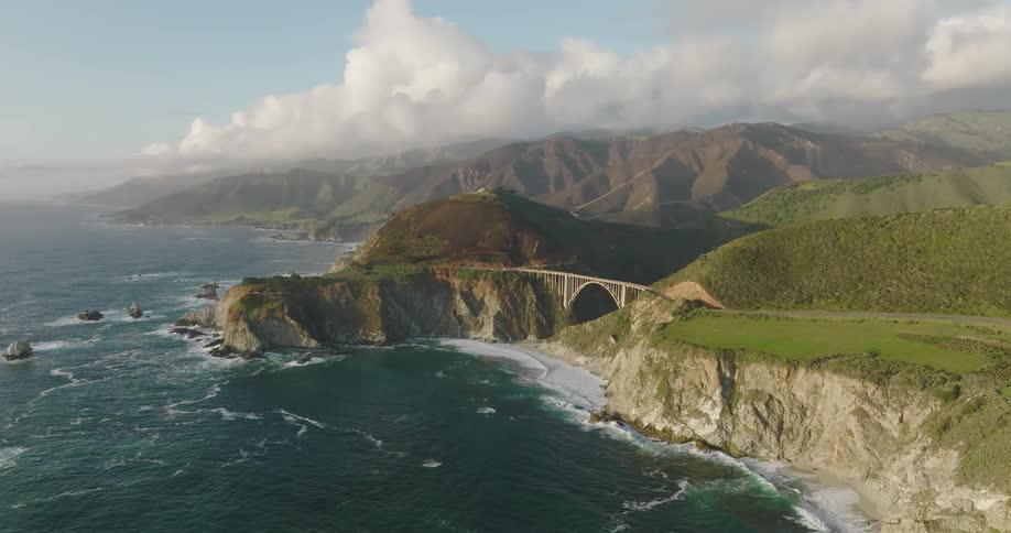 Waves crash against cliff base near bridge on foggy day