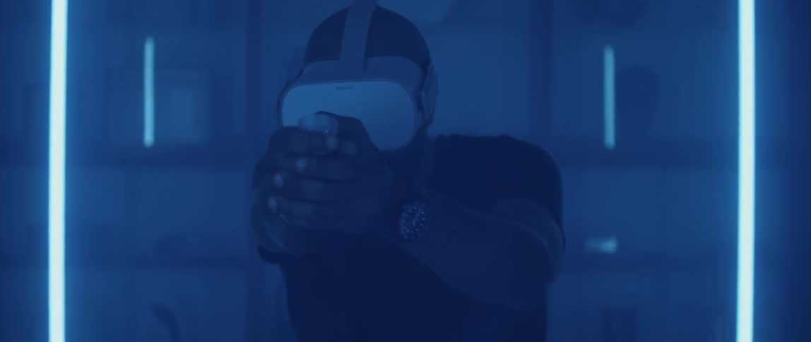A man makes gun firing motions while playing a game on a virtual reality headset in a dimly lit room