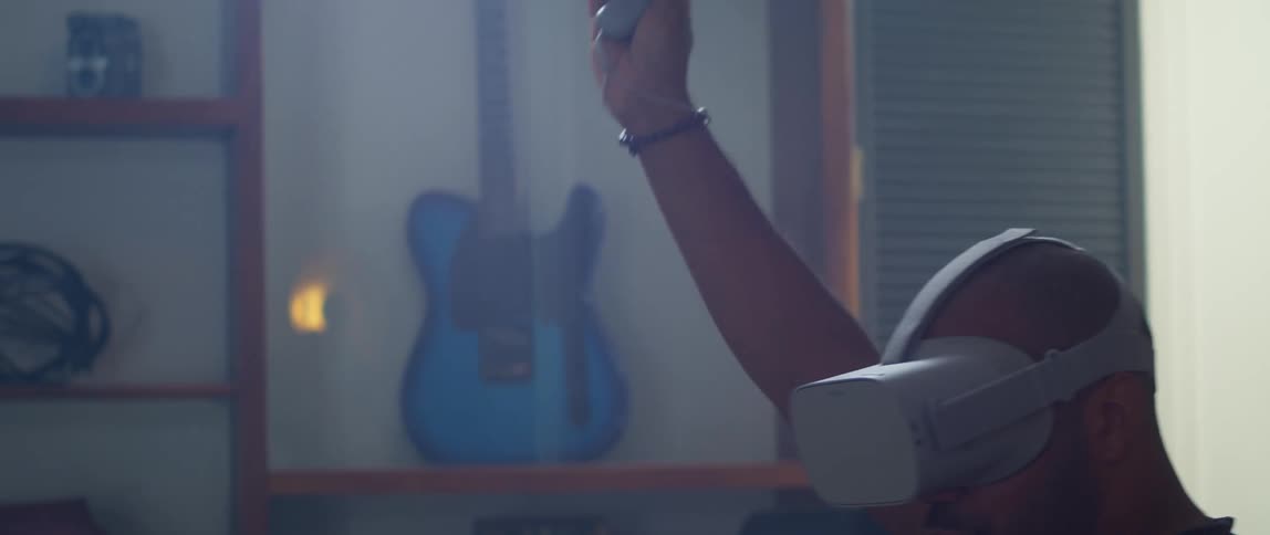 A man is sitting on a living room couch wearing a virtual reality headset and moves his arm with a remote controller in his hand as he plays