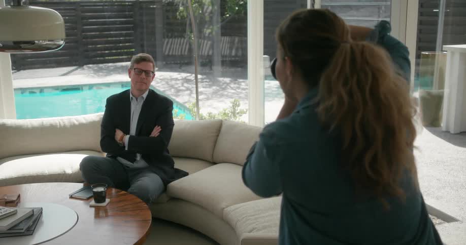 A man in a suit is sitting on a couch and a woman is taking his picture