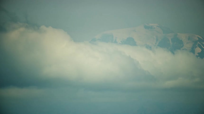 Cumulus clouds move across the sky with snow covered mountains in the distance from this view out the window of a passenger jet liner