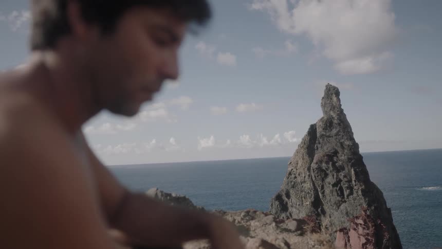 A man who is looking down turns his head to look towards a rock and ocean in the distance