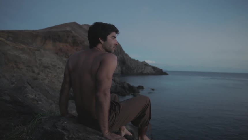 A shirtless man sits on a hilly coastline