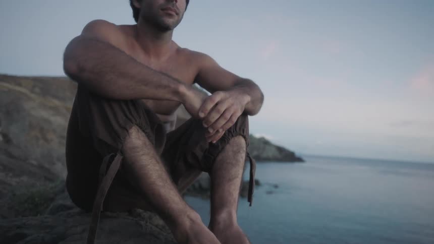 A shirtless man sits on a rock while overlooking the ocean at dusk