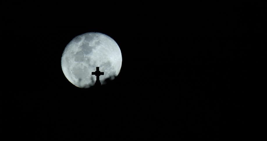 The full moon rises over a cemetery hill behind a cross shaped gravestone in the darkness