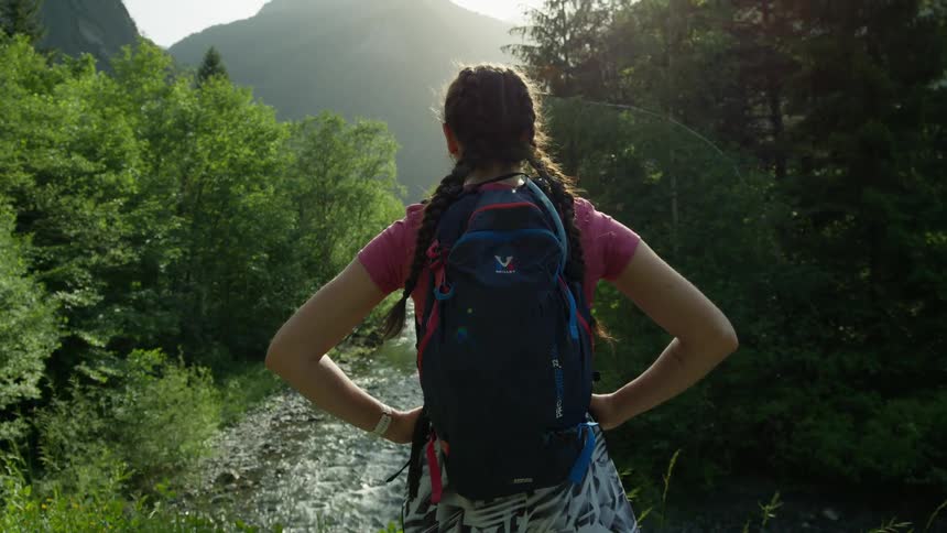 A woman wearing a backpack is standing in the woods with her hands on her hips overlooking a stream as the sun shines brightly over the mountains in the distance