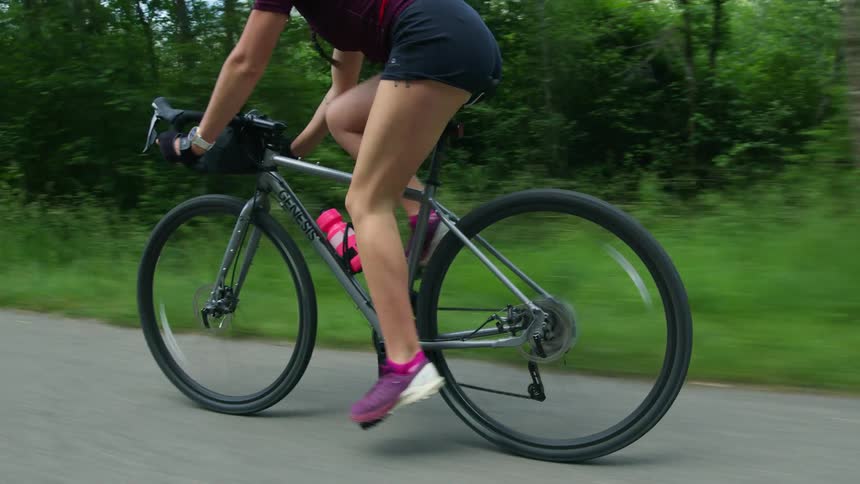 A woman is riding her bicycle near the woods and she has a pink water bottle on her bike