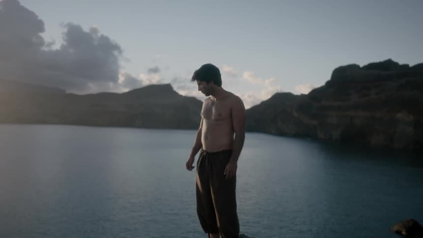A shirtless man stands on a rock overlooking a body of water