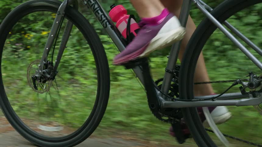 A woman is pedaling a bicycle near the woods and she has a pink water bottle on her bike