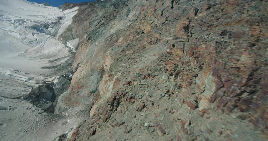 Steep bare rock mountain side abuts snow covered slope under a clear blue sky