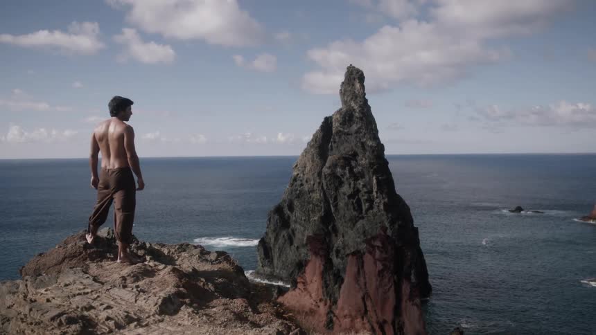 Shirtless man standing on a high cliff looking out at the ocean