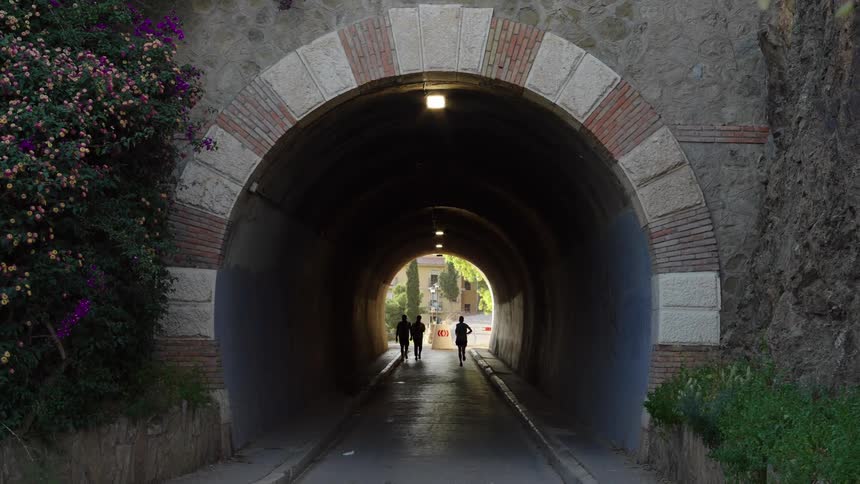 Pedestrians and jogger moving through tunnel are silhouetted against buildings on the other side
