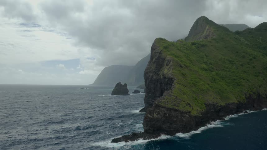 Waves splash against rock formations and mountains along the coast under a cloudy sky