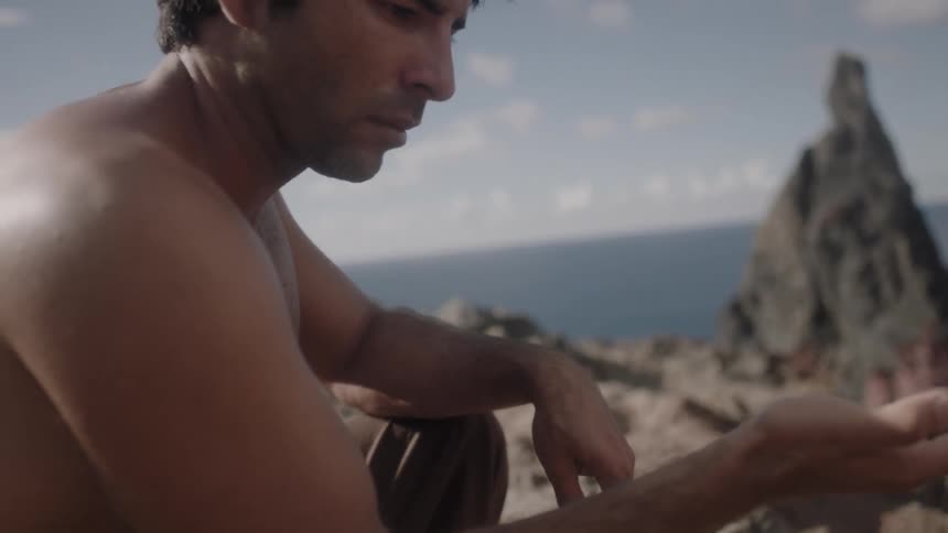 A man perched on a cliff above a shoreline is staring intently at something in his outstretched hand