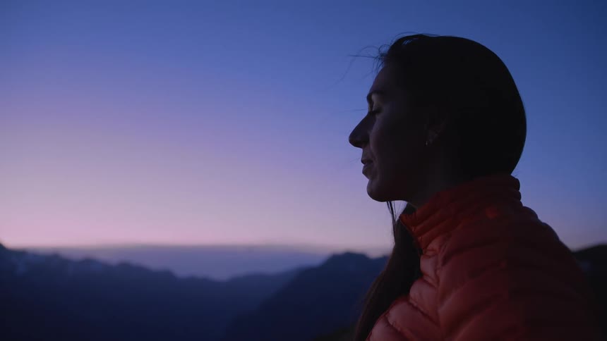 Woman in orange coat looks into space on mountain at dusk