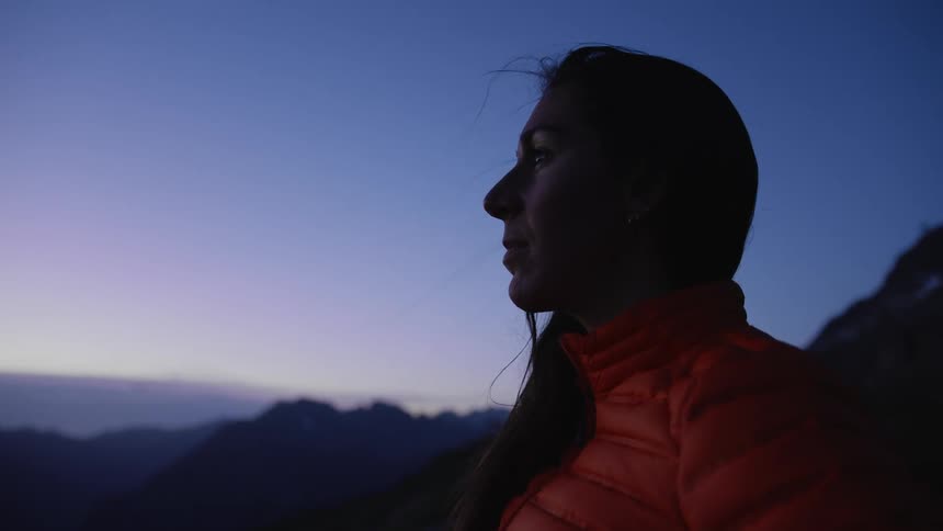 A woman is standing outdoors and wearing a jacket at dusk
