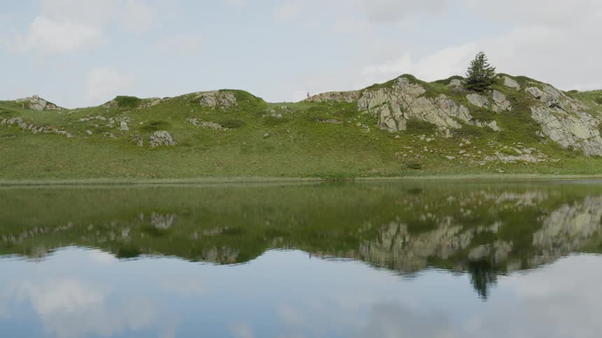 A jogger is running on a path across the top of a grass covered mountain as the reflection shines in the lake below