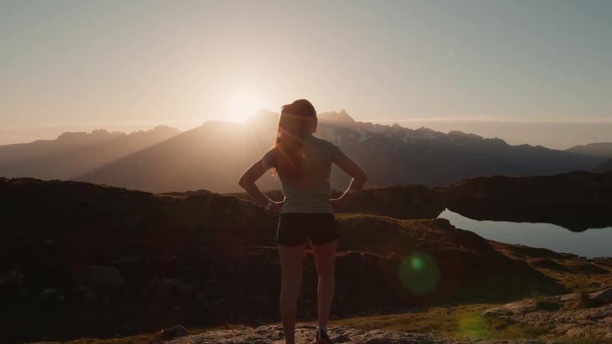 A woman is standing outside on a ledge looking at mountains as the sun shines brightly over the peaks