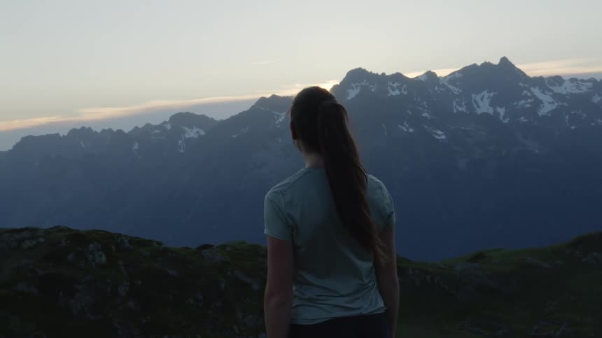 A woman is standing outside on a ledge looking at mountains with peaks high in the sky