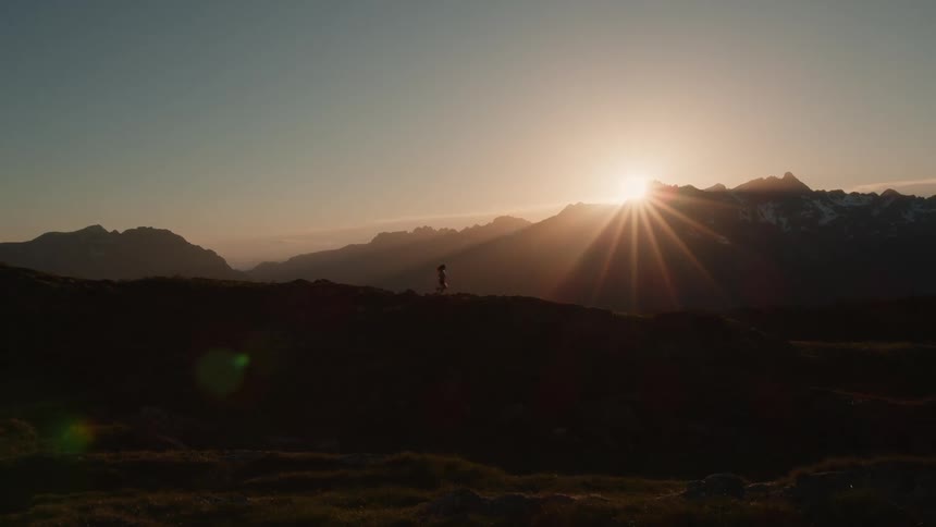 Woman runs across hill top silhouetted by the sun cresting nearby mountains