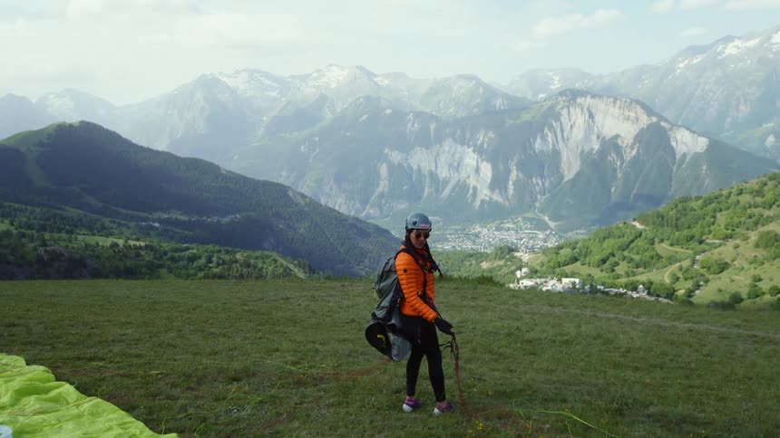 A young white woman is standing on a hill in front of a huge mountain getting ready to paraglide