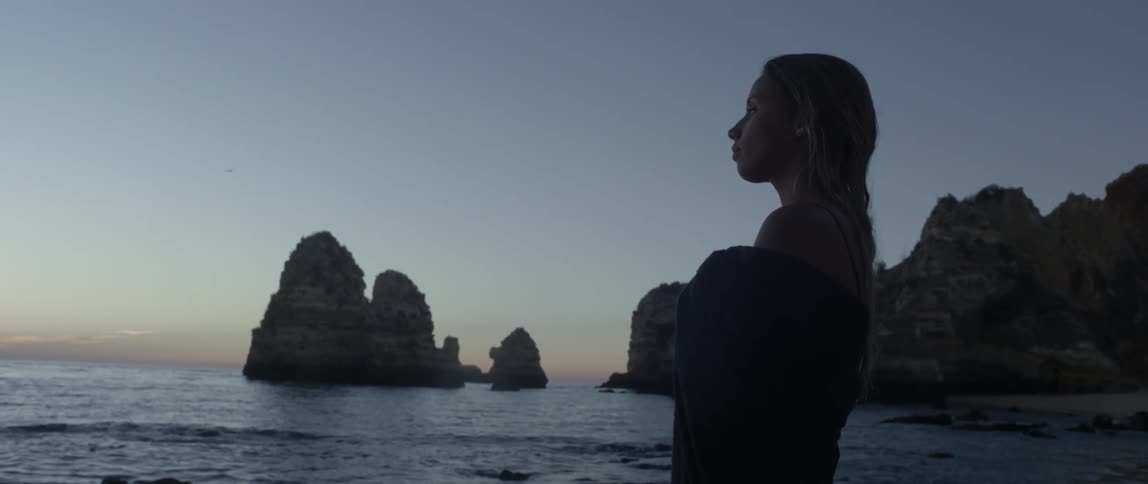 A woman stands on a beach while looking out at the ocean at dusk