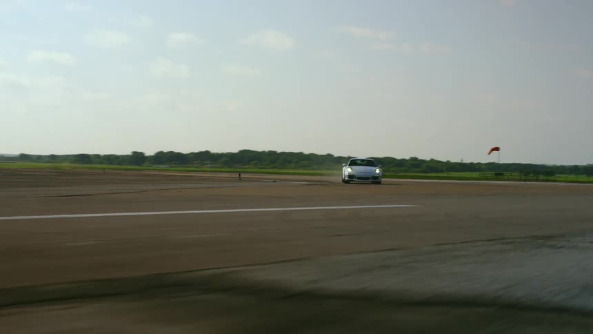 Car speeds along wide course bordered by fields with mountains and forest on horizon