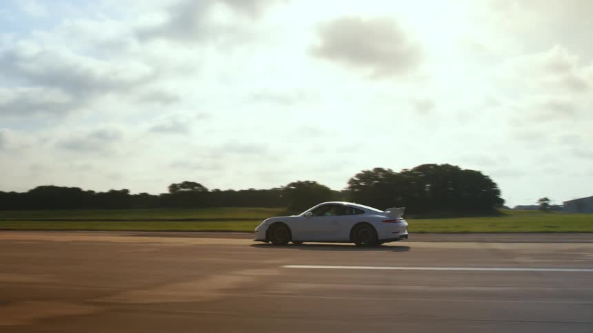 A person driving a porche 911 gt3 on an airport runway with grass and trees in the distance