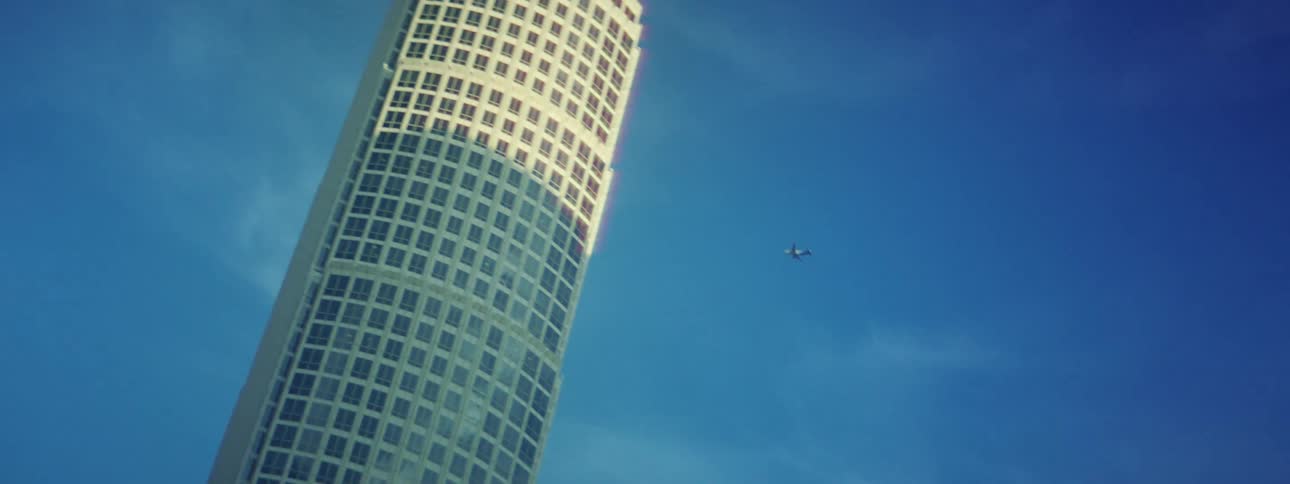 An airplane flies overhead of a towering building on a sunny day