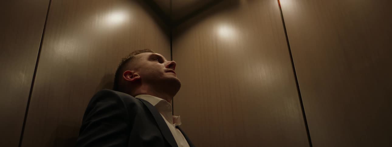 A man leaning against an elevator wall slides to the corner of the car