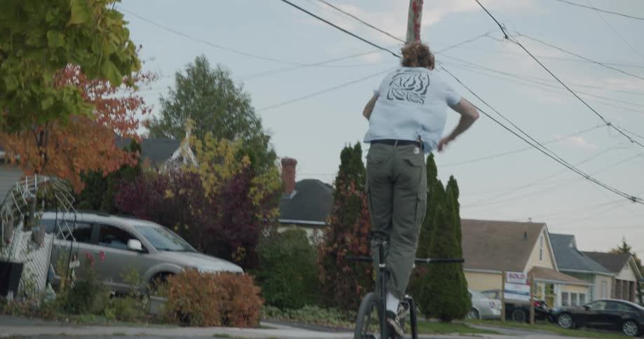 Man wearing white shirt and green pants does tricks on bike down the street in the neighborhood