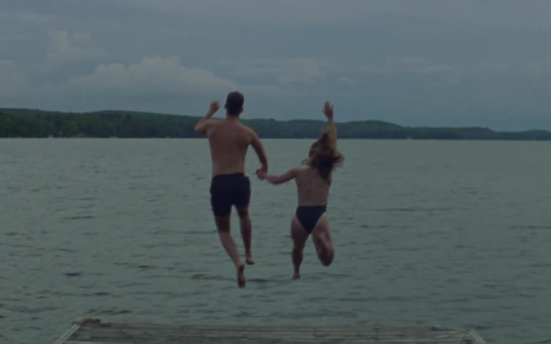 A couple holding hands runs down a dock and jumps into a lake