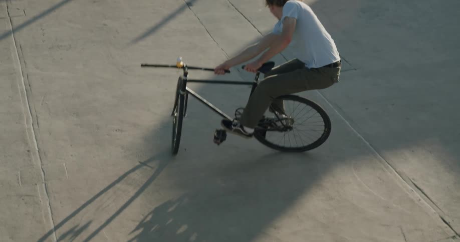 Man with long hair white shirt and green pants does trick on bicycle in open public space