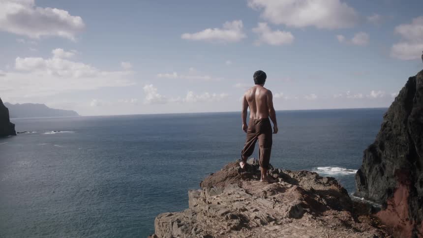 A man stands on the edge of a cliff by the shore