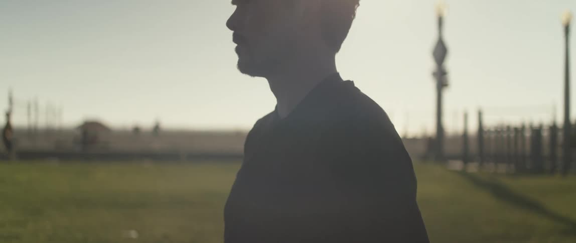 A man is stretching his legs in a field in a park as people are walking and riding bicycles in the distance