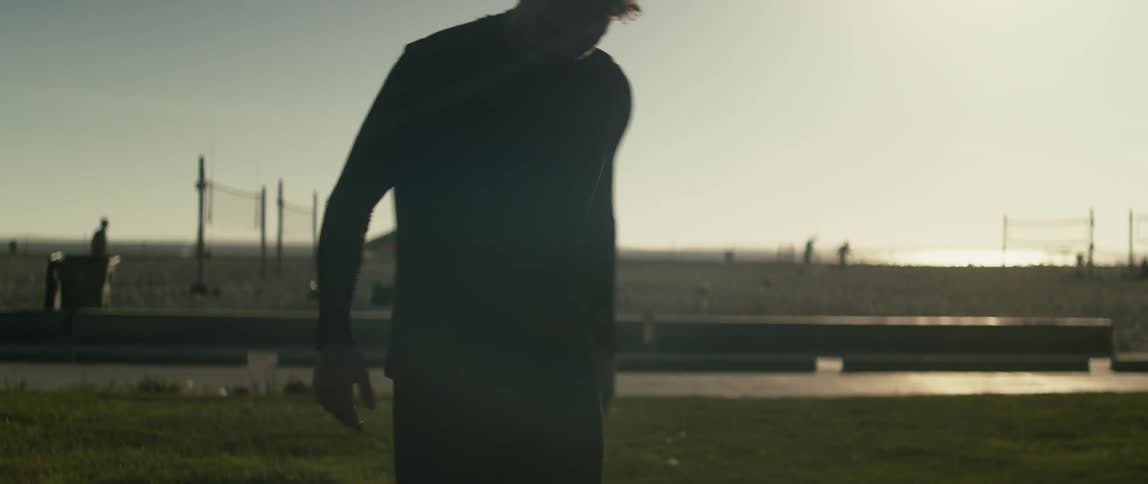 A teenager kicks a soccer ball as people behind him roller skate walk and jog down a path