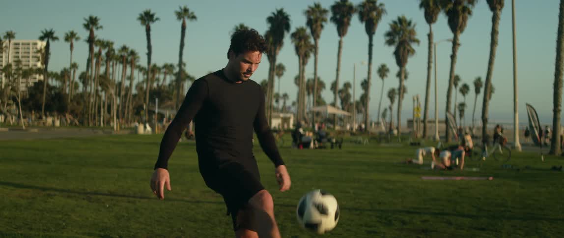 A man kicking a soccer ball in a grassy field with palm trees in the distance