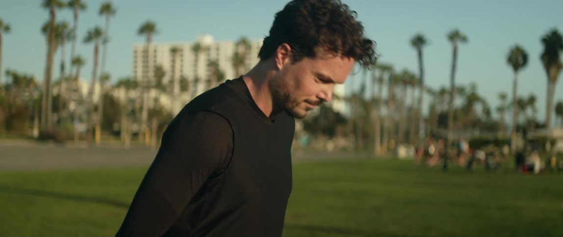 A man kicks a ball in a public park with many people gathering on the grass in the background