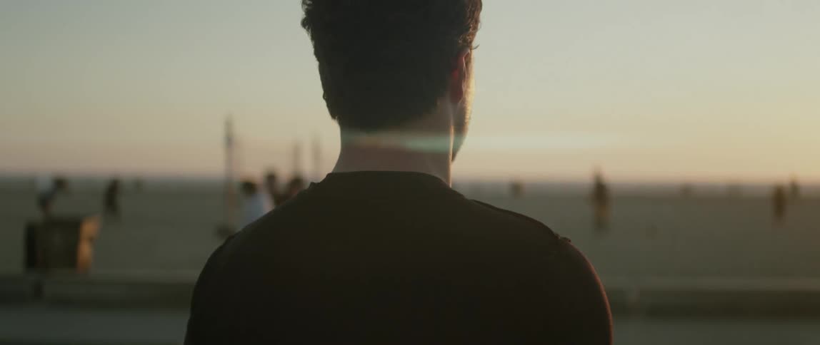A man is standing in a park holding a soccer ball and watching people walking and playing on the beach as the sun shines brightly on the horizon