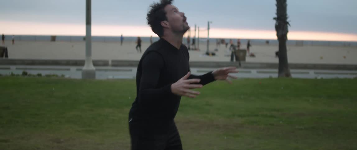 A man is standing in a field and he tosses a soccer ball into the air and catches it on the back of his neck