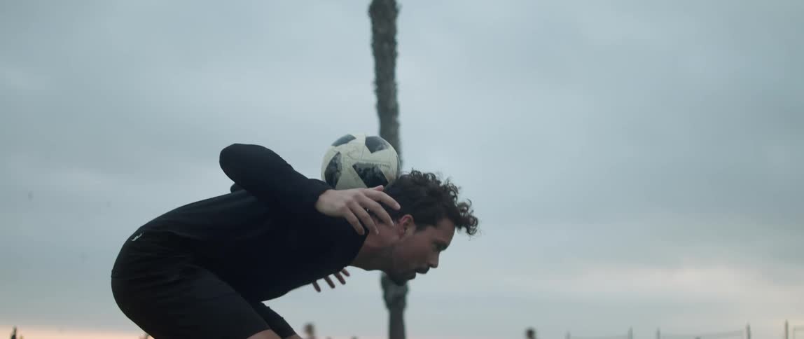 Man wearing black sweatshirt and shorts does tricks with soccer ball near the beach