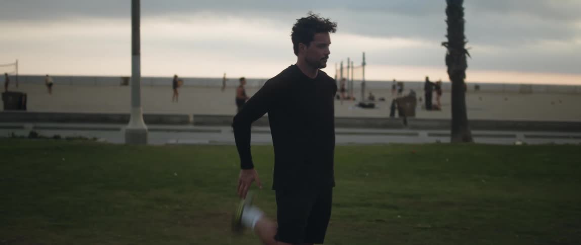 Man stretches his leg behind him while standing in park across from busy beach