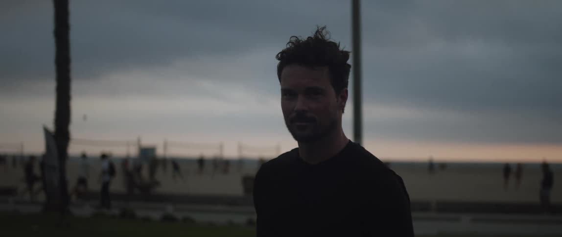 A man is walking outside as many people are walking and playing on the beach in the distance