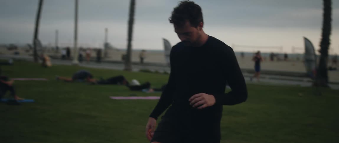 A man at a public park by a beach kicks a soccer ball as other people lie on the grass ride bikes jog and play volleyball