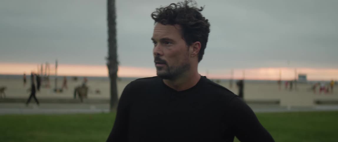 A man holding a soccer ball in front of a beach filled with people looks off in to the distance and then turns around