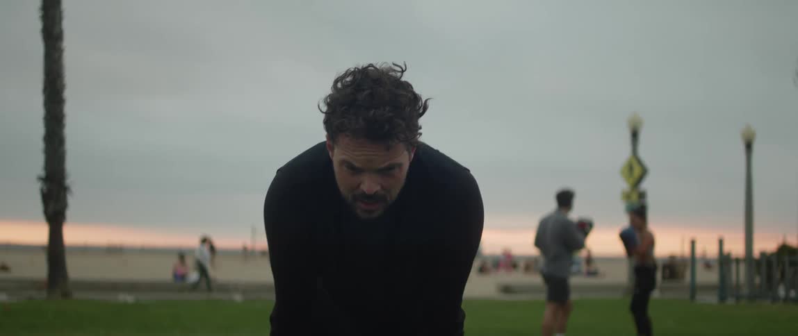 A man is standing in a field bending down with his hands on his legs and takes a deep breath as people are walking and playing in the distance