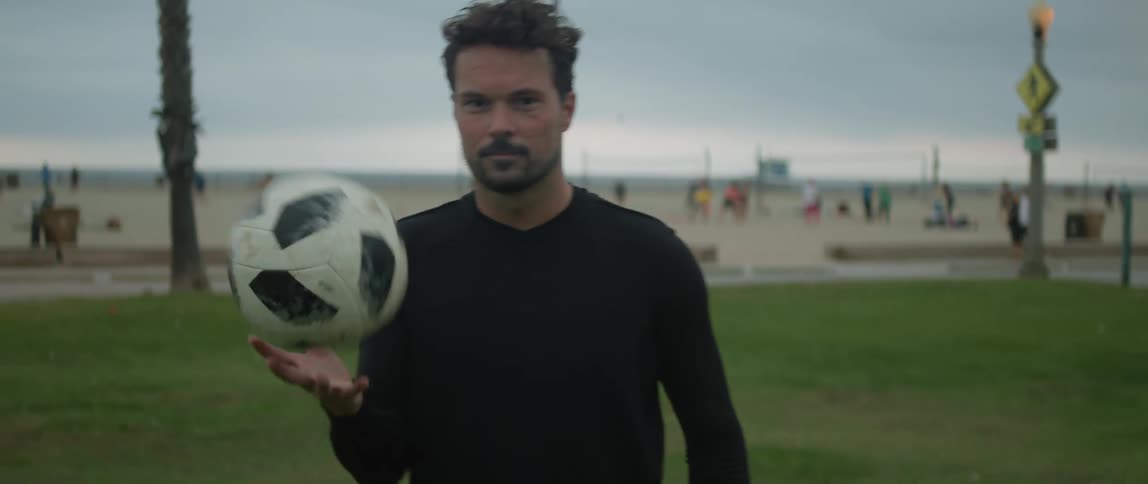 Man with black sweatshirt and shorts juggles a soccer ball on green lawn next to the beach
