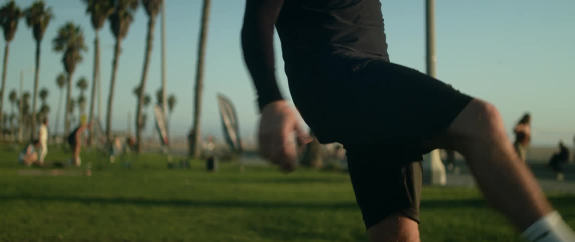 A man is bouncing a soccer ball into the air using his feet and knees as people walk in the distance