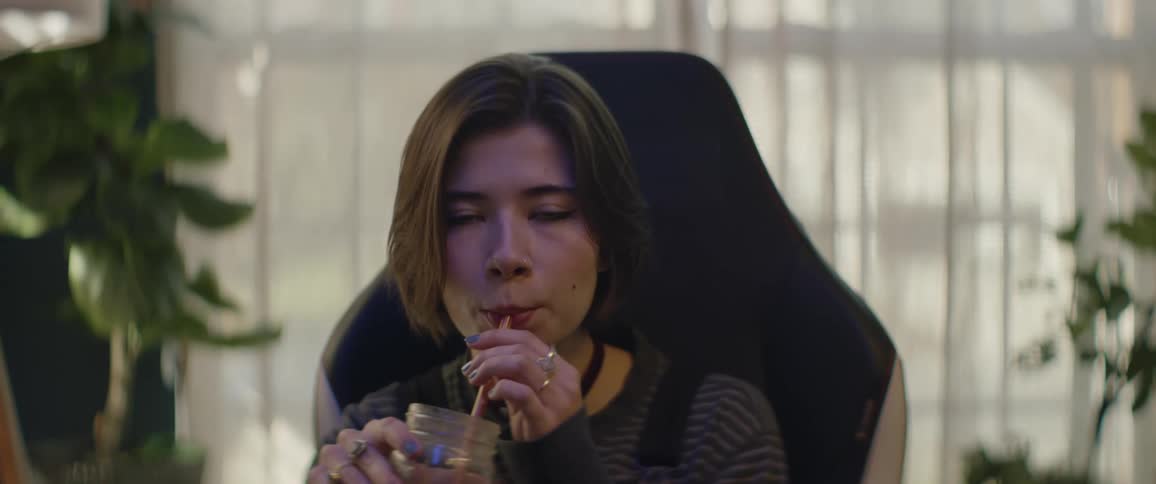 A young woman sitting indoors uses a straw to drink from a jar while looking into the camera